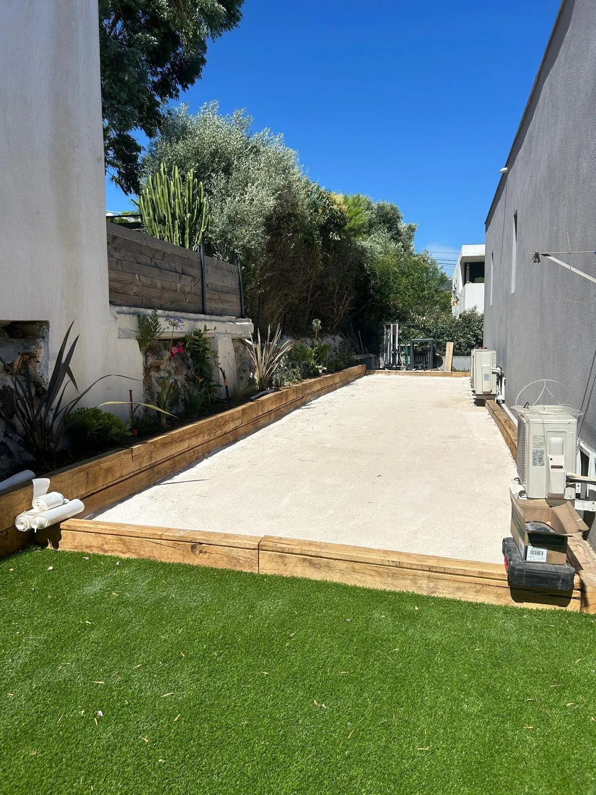 Terrain de pétanque entre un mur et un muret planté, gazon au premier plan