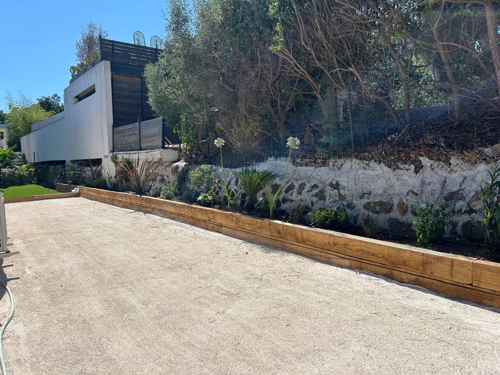 Terrain de pétanque bordé de traverses en bois, au pied d’un muret planté